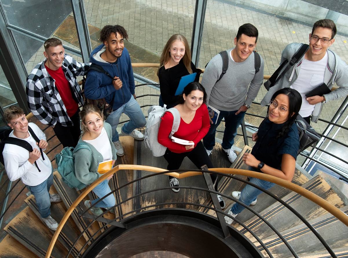 9 Studierende stehen gemeinsam auf einer Treppe und schauen lachend in die Kamera.