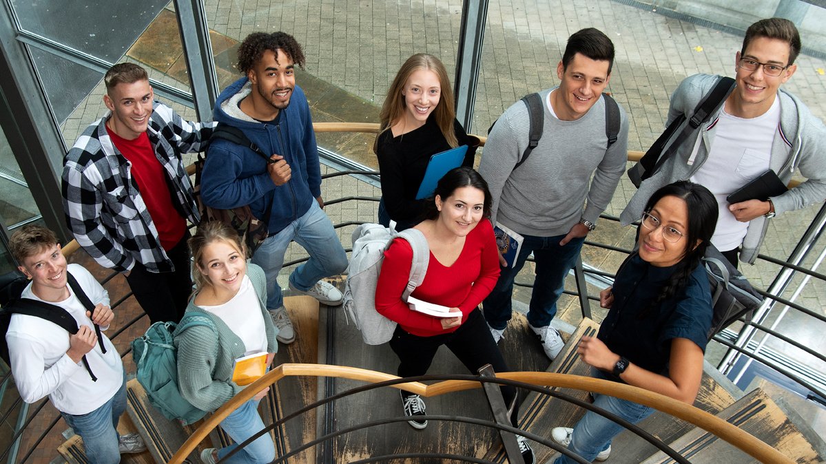 9 students stand together on a staircase and look into the camera laughing.